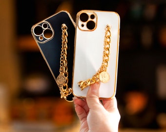 Puede incluir: Dos fundas para teléfono, una negra y otra blanca, con bordes dorados y una correa de cadena dorada. La cadena tiene un dije de moneda. Las fundas están diseñadas para proteger el teléfono y agregar un toque de elegancia.