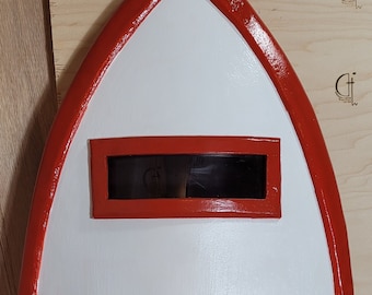May include: An oval shield, painted white with a red border and a rectangular black viewing aperture. The shield's shape is elongated, and it appears to be made of wood or a similar material.