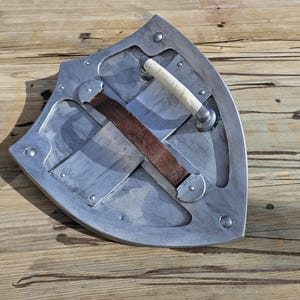 May include: A silver-colored shield with a curved design, featuring a white handle and a brown leather strap. The shield is mounted with rivets and has a worn appearance, suggesting a medieval or fantasy theme.