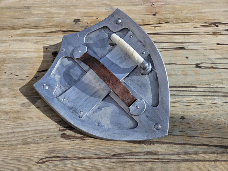 May include: A silver-colored shield with a curved design, featuring a white handle and a brown leather strap. The shield is mounted with rivets and has a worn appearance, suggesting a medieval or fantasy theme.