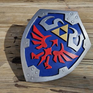 May include: A blue, silver, and red shield with a gold triangle in the center. The shield has a silver border with rivets and a red bird-like emblem. The shield is a prop from a video game.