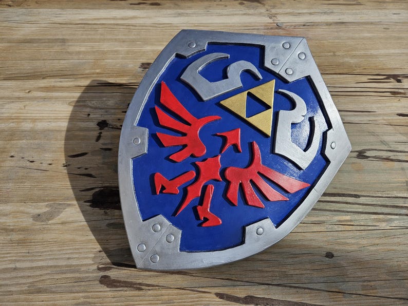 May include: A blue, silver, and red shield with a gold triangle in the center. The shield has a silver border with rivets and a red bird-like emblem. The shield is a prop from a video game.