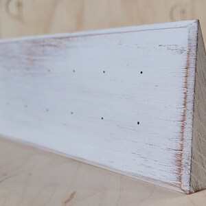 May include: A white, wooden earring display stand with a distressed finish. The stand is triangular, with a flat front surface featuring several small holes for earrings. The wood grain is visible, adding a rustic touch.