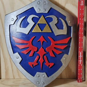 May include: A blue, silver, and red shield with a gold triangle in the center. The shield has a stylized design with red wings and silver accents. The shield is a prop from a video game.