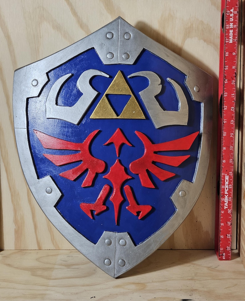 May include: A blue, silver, and red shield with a gold triangle in the center. The shield has a stylized design with red wings and silver accents. The shield is a prop from a video game.