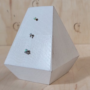 Może przedstawiać: Biała, geometryczna rzeźba z drewna o kształcie rombu. Trzy małe, dekoracyjne, opalizujące ćwieki są przymocowane do przodu. Rzeźba jest ustawiona na jasnym tle drewnianym.