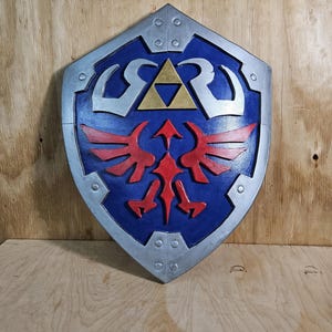 May include: A blue and silver shield with a red emblem and a gold triangle in the center. The shield has a silver border and is set against a wooden background. The shield is a prop from the Legend of Zelda.