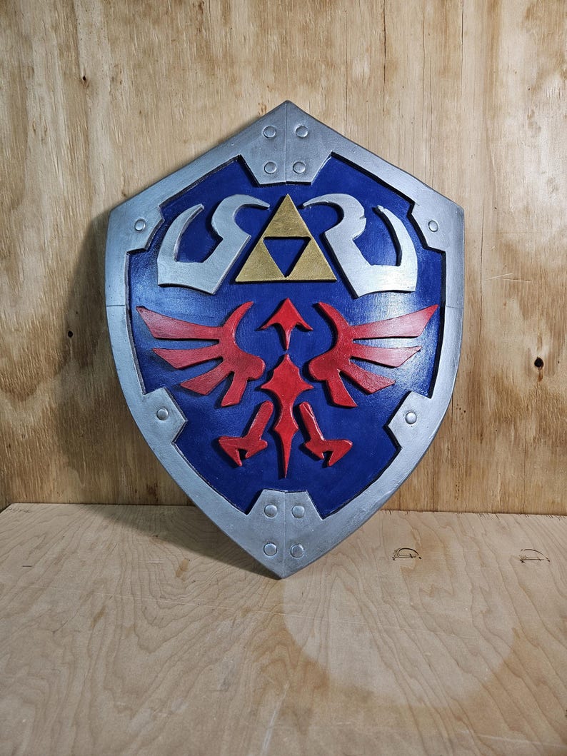 May include: A blue and silver shield with a red emblem and a gold triangle in the center. The shield has a silver border and is set against a wooden background. The shield is a prop from the Legend of Zelda.
