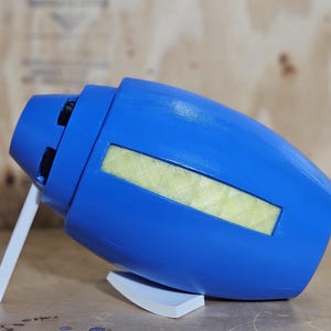 May include: A blue prop resembling a futuristic weapon, with a cylindrical body and a rectangular yellow panel. It has a white support stand and a white angled support. The prop is likely a cosplay accessory or a decorative item.