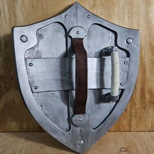 May include: A silver-colored shield with a pointed top and rounded sides. It features a brown leather strap and a white-wrapped handle on the back. Rivets and metal plates are visible on the shield's surface. The shield is set against a wooden background.