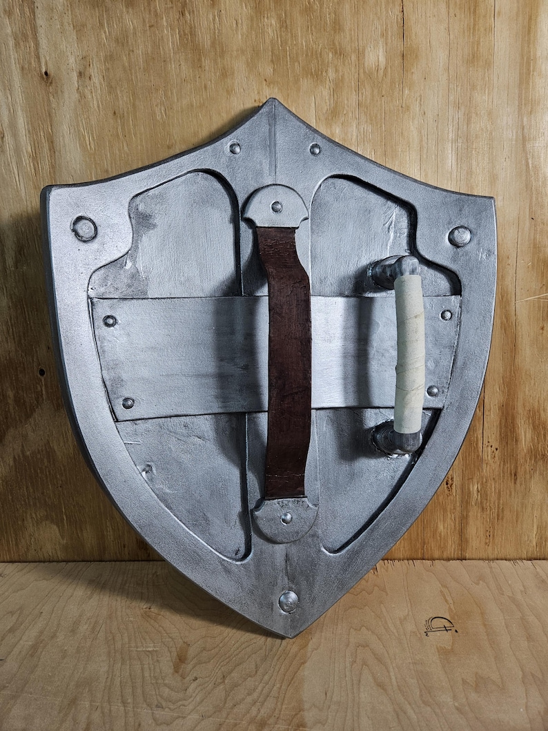 May include: A silver-colored shield with a pointed top and rounded sides. It features a brown leather strap and a white-wrapped handle on the back. Rivets and metal plates are visible on the shield's surface. The shield is set against a wooden background.