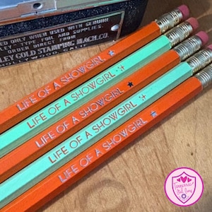 May include: A close-up shot of five pencils with the text "LIFE OF A SHOWGIRL" in white. The pencils alternate between orange and teal, with pink erasers. The pencils are on a wooden surface.