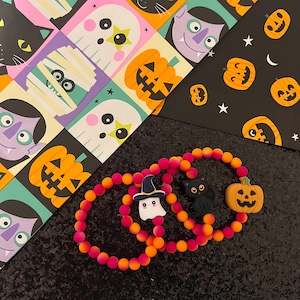 May include: Halloween-themed beaded bracelets in shades of pink, orange, and yellow. Each bracelet features a charm, including a black cat, a jack-o'-lantern, and a ghost wearing a witch hat. The background includes Halloween-themed patterned paper.