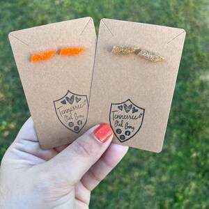 May include: Two pairs of earrings on brown card stock. The earrings are shaped like the state of Tennessee. One pair is orange glitter and the other is gold glitter. The card stock has the text "Tennessee Girl Gang" with social media icons.