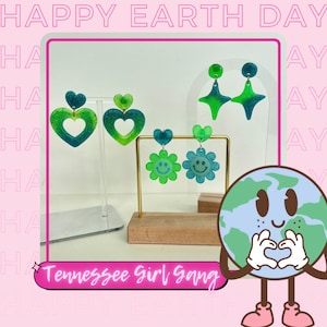 May include: Green, blue, and white resin earrings in the shapes of hearts, stars, and flowers. The earrings are displayed on a wooden stand with a cartoon Earth holding a heart in its hands. The text "Tennessee Girl Gang" is visible in the image.