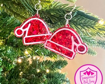 May include: Red glitter Santa hat earrings with white outlines. The earrings are hanging from a Christmas tree.