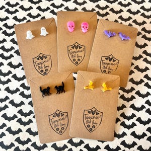 May include: A collection of Halloween-themed stud earrings, including ghosts, skulls, bats, black cats, and witches on brown cards. Each card has a logo that reads "Tennessee Girl Bang". The earrings are in various colors, including white, pink, purple, black, and yellow.