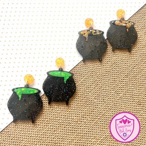May include: Three pairs of black cauldron earrings with glitter. One pair has green slime dripping from the cauldron, one pair has orange and black sprinkles, and one pair has orange and yellow sprinkles.