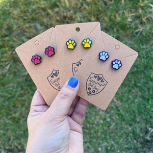 May include: Three pairs of paw print earrings on brown cards. The earrings are red, yellow, and blue with black glitter. The cards have the text "Tennessee Girl Gang" on them.