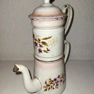 以下が含まれることがあります： ピンクと花の模様が施された白いエナメル製のコーヒーポット。ポットは3段構造で、上に蓋が付いています。