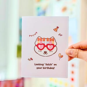 May include: A white birthday card featuring a cartoon dog with heart-shaped glasses and a party hat. The card reads "Looking 'fetch' on your birthday!" and is decorated with confetti and balloons.