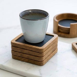 Puede incluir: Un juego de posavasos cuadrados de madera con inserciones negras, con una taza de cerámica azul claro encima. También se ven posavasos adicionales y un soporte. Los posavasos son de madera y tienen un centro oscuro, casi negro.