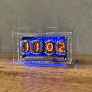 Puede incluir: Una caja de acrílico transparente con una luz azul ilumina cuatro tubos Nixie que muestran el número 1102.
