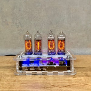 Puede incluir: Una caja de acrílico transparente con cuatro tubos Nixie naranjas brillantes que muestran el número "1000". Los tubos están iluminados con un tono púrpura. La caja tiene una pequeña placa de circuito visible en el interior.