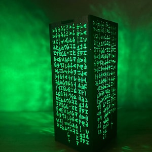 May include: A black wooden box with a green light shining through it. The box is covered in a pattern of symbols and letters. The box is a decorative lamp.