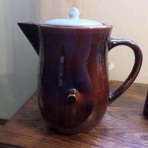 May include: A brown ceramic coffee pot with a light blue lid. The pot has a handle and a spout.