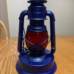 May include: A blue metal lantern with a red glass globe. The lantern has a handle at the top and the words "LITTLE GIANT" are printed on the base. The lantern is sitting on a wooden table.