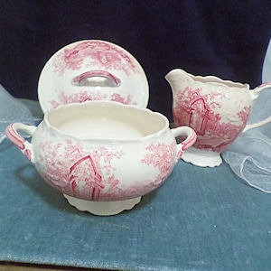 May include: A vintage porcelain sugar bowl and creamer set with a pink floral design. The set features a lidded sugar bowl and a creamer with a handle.