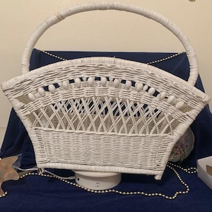 May include: A white wicker basket with a curved handle. The basket has a woven design and is decorated with small, white, oval-shaped accents along the top edge. The basket sits on a white base and is set against a dark blue background.
