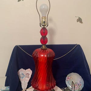 May include: A red glass table lamp with a brass base and a clear glass shade. The lamp has a ribbed design and a round, red glass finial.