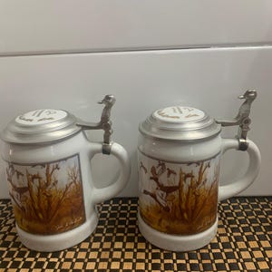 May include: Two white ceramic beer steins with lids. Each stein features a handle, a metal lid, and a decorative scene of ducks in a marsh. The lids have a duck-shaped lever. The steins are on a brown and black checkered surface.