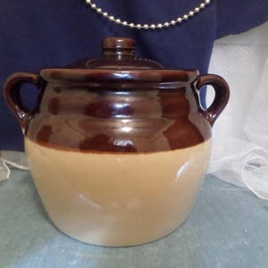 May include: A brown and beige ceramic crock with two handles and a lid. The crock is glazed and has a rustic look.