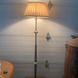 May include: A floor lamp with a tall, slender, twisted metal stand featuring a dark finish and a small, round, light-colored stone accent. The lamp is topped with a conical, beige-colored fabric lampshade with vertical stripes and a warm glow.
