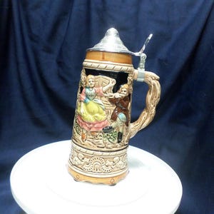 May include: A ceramic beer stein with a brown and tan glaze. The stein features a scene of a man and woman dancing. The stein has a metal lid with a hinged handle.