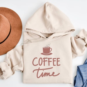 May include: A beige hoodie with a coffee cup graphic and the words "COFFEE time" in a script font. A brown hat is visible in the upper left corner. The hoodie has a drawstring and a kangaroo pocket.
