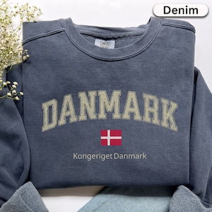 May include: Denim blue sweatshirt featuring the word "DANMARK" in a vintage font, a Danish flag, and the text "Kongeriget Danmark". The sweatshirt is folded on a light blue denim surface.