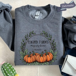 以下が含まれることがあります： オレンジ色のカボチャと緑の葉の円形デザインで「Hagrid Farms Magically Grown Pumpkins」の文字が入ったダークヘザーグレーのスウェットシャツ。スウェットシャツはクルーネックです。