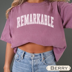 Peut inclure: Un t-shirt de couleur baie avec le mot "REMARKABLE" imprimé en lettres majuscules blanches sur la poitrine. Le t-shirt a des manches courtes et une coupe décontractée. Le modèle porte un jean en denim bleu clair.