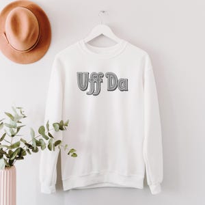 Peut inclure: Sweat-shirt blanc avec le texte "Uff Da" en gris ombré. Le sweat-shirt est suspendu à un cintre blanc. Un chapeau marron et un vase avec des feuilles vertes sont visibles en arrière-plan.