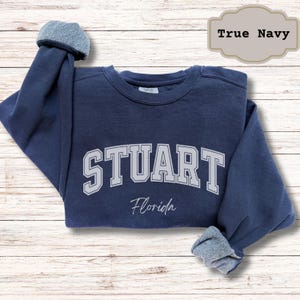 May include: A navy blue sweatshirt with the word "STUART" in white block letters, with "Florida" in script below. The sleeves are cuffed, revealing a lighter blue lining. The text "True Navy" is in a light brown label.