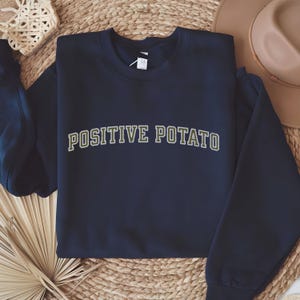 May include: Navy blue sweatshirt with the text "POSITIVE POTATO" in a green and white arched design. The sweatshirt is made of a soft material and is suitable for casual wear.