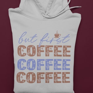 Może przedstawiać: Biała bluza z kapturem z napisem "but first COFFEE" w niebieskich i brązowych kwiatowych literach. Mała grafika filiżanki do kawy znajduje się nad słowem "coffee". Bluza z kapturem ma kaptur ze sznurkiem i jest złożona na bordowej powierzchni.