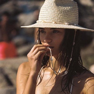 Può includere: Un cappello di paglia con dettagli di conchiglie e un top bikini a righe verdi e bianche. Il cappello ha un'ampia tesa ed è decorato con piccole conchiglie. È visibile anche una collana con conchiglie.