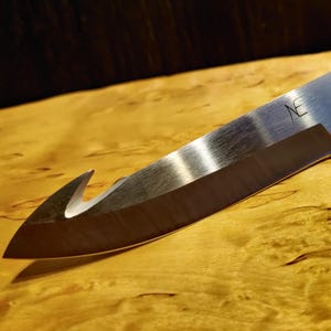 Puede incluir: Un cuchillo plateado con una hoja curva y un gancho en la punta. La hoja tiene un borde dentado y un pequeño agujero cerca del mango. El cuchillo está sobre una superficie de madera.