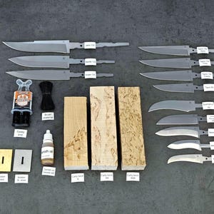 Op de afbeelding: Een verzameling materialen voor het maken van messen, waaronder onafgewerkte mesbladen, houten handvatblokken, epoxy, afwerkingsolie en een borstel. De bladen zijn zilverkleurig en het hout vertoont verschillende patronen.