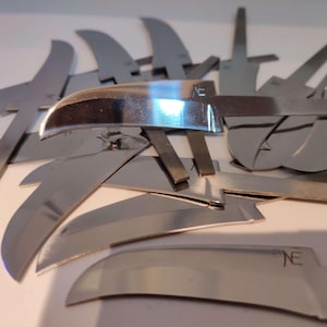 May include: A close-up of a pile of silver knife blades, some of which have the letter "NE" engraved on them.